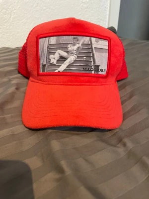 Röd truckerkeps från Maggiore - Snygg röd truckerkeps från Maggiore med mesh på sidorna och baksidan. Framtill finns en svartvit bild med tryck och texten 'MAGGIORE'. Kepsen har böjd skärm och justerbar passform. Perfekt för dig som vill sticka ut med en unik accessoar.