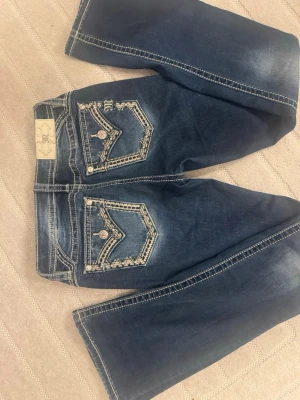 Mörkblå jeans med dekorerade fickor - Säljer ett par miss me jeans. Dom är mörkblå jeans med snygga slitningar och detaljerade bakfickor med broderier och paljetter. Jeansen är bootcut. Dom är storlek 26. 
