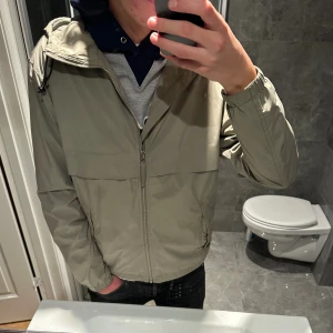 Olivgrön vindjacka med huva  - Säljer en vindjacka u riktigt snygg olivgrön färg. Jackan är i toppskick och saknar spår av användning. Jag på bilden är 188cm och den är något kort på mig. Hör av er vid funderingar:)