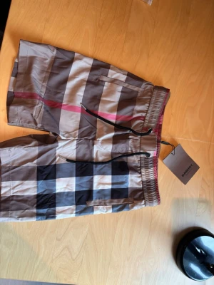 Burberry shorts storlek S - Säljer ett par klassiska rutig shorts från Burberry med snörning i midjan och elastisk linning. Shortsen har stora rutor i beige, svart, vit och röd, samt fickor på sidorna. Perfekta för sommaren och riktigt snygga till streetwear-stilen.