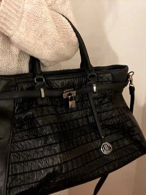Svart handväska  - Snygg svart handväska i fejkläder med mönster. Väskan har dubbla handtag, avtagbar axelrem, silverfärgade detaljer. Perfekt för dig som vill ha en trendig och rymlig väska för skolan eller shopping!