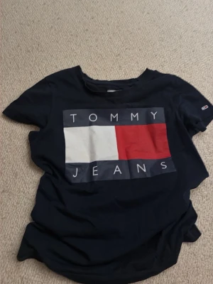 Marinblå Tommy Jeans t-shirt - Säljer en marinblå t-shirt från Tommy Jeans med stort tryck på bröstet i vitt, rött och blått. Klassisk passform och rund hals. Perfekt för dig som gillar stilrena logotishirts.