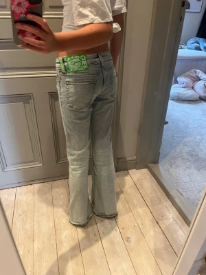 Ljusblå bootcut jeans med låg midja - Säljer ett par ljusblå bootcut jeans med låg midja och klassiska fem fickor. Jeansen har en snygg tvätt med slitningar framtill och en grön detalj på linningen bak. Perfekta för en trendig och avslappnad look.