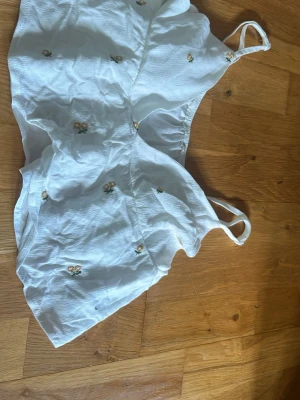Vit blommig linnetopp från SHEIN - Söt vit linnetopp från SHEIN med tunna axelband och små broderade blommor i gult och grönt. Toppen har en lätt och luftig känsla, perfekt för varma dagar. Passar dig som gillar en feminin och somrig stil.