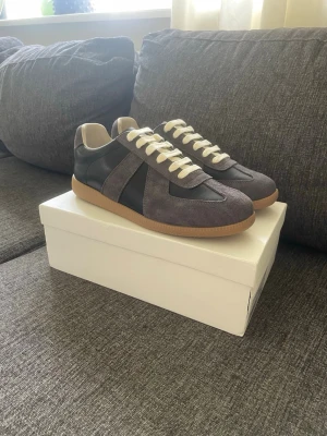 Maison Margiela Gats - Snygga sneakers i grå mocka och svart läder med vita snören och beige innersula. Skorna har en klassisk låg siluett och gummisula i ljusbrunt. Perfekta för dig som gillar stilrena och tidlösa sneakers.