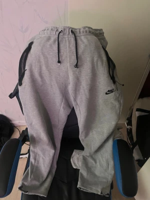 Gråa mjukisbyxor från Nike - Snygga grå mjukisbyxor från Nike med svarta detaljer och dragsko i midjan. Byxorna har två sidofickor med dragkedja och Nike-logga på vänster ben. Perfekta för chill dagar eller träning.