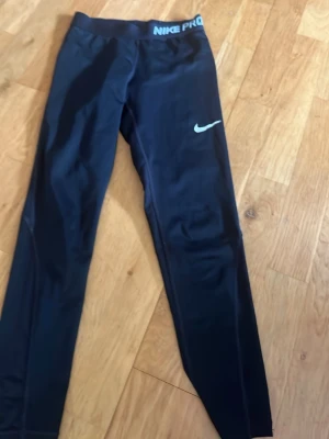 Svarta Nike Pro leggings - Snygga svarta Nike Pro leggings med vit logga på benet och Nike Pro-tryck i midjan. Tight passform och stretchigt material som passar perfekt till träning eller chill. Klassisk design med hög midja och långa ben.