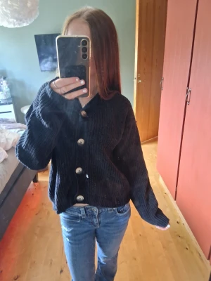 Svart stickad kofta med stora knappar - Mysig svart stickad kofta med oversized passform och stora, kontrasterande knappar framtill. Koftan har långa ärmar och ribbade muddar vid ärmslut och nederkant. Perfekt att slänga över en t-shirt för en avslappnad look.💕