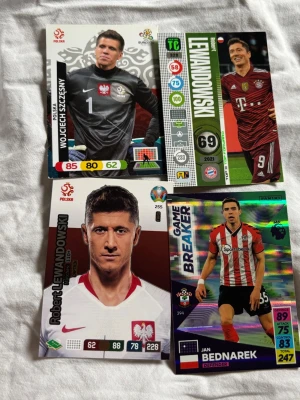 Fyra fotbollssamlarspelkort - Säljer fyra samlarkort med fotbollstema från olika spel, med spelare som Robert Lewandowski, Wojciech Szczęsny och Jan Bednarek. Korten har färgstarka designer, olika statistik och klubb- eller landslagsloggor. Perfekt för dig som gillar fotboll och samlarkort.