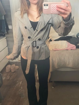 Grå rutig trenchcoat med bälte - Snygg grå trenchcoat med diskret rutmönster och svarta knappar. Jackan har klassisk krage, axelklaffar och tillhörande bälte i midjan för en markerad siluett. Perfekt för dig som gillar stilrena och trendiga ytterplagg.