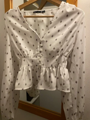 Vit blus med brunt mönster och knappar - Söt vit blus från Lager 157 med brunt småblommigt mönster, v-ringning och knappar framtill. Blusen har lång ärm och volang nedtill i midjan. Passar xs/s💕