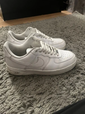 Nike Air Force 1 sneakers  - Klassiska helvita Nike Air Force 1 sneakers i lågt utförande. Skorna har en stilren design med perforerad tåbox, vit sula och ikoniska swoosh-loggan på sidan. Tillverkade i läder med matchande vita skosnören och detaljer.