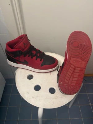 Svarta och röda Air Jordan 1 Mid - Säljer ett par Air Jordan 1 Mid i svart och rött med klassisk design och svart swoosh. Skorna har en vit mellansula och röd yttersula, samt ikoniska Jordan-loggor på plösen och sidan. Perfekta för dig som gillar streetwear och sneakerskultur.