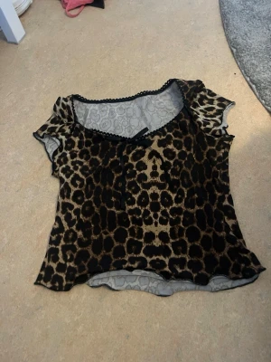 Leopardmönstrad mesh topp med rosett - Snygg leopardmönstrad mesh topp med korta ärmar och liten svart rosett vid urringningen. Toppen har vågig kant och transparent material som ger en trendig look. Perfekt för dig som vill sticka ut lite extra.