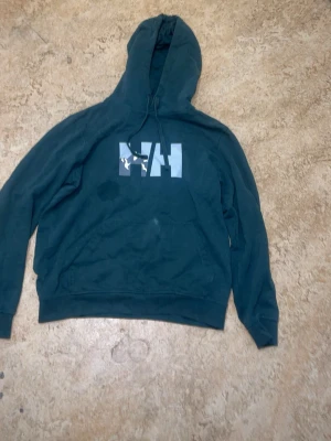 Mörkgrön hoodie från Helly Hansen - Mörkgrön hoodie från Helly Hansen med stor vit HH-logga på bröstet. Klassisk huva med dragsko och ribbade muddar vid ärmslut och nederkant. Tillverkad i mjukt bomullsmaterial, perfekt för chill dagar eller streetwear-stil.