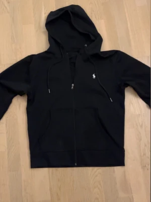 Svart hoodie med dragkedja - Svart hoodie med dragkedja och huva, samt dragsnören och en liten vit logga på bröstet. Klassisk känguruficka framtill och ribbade muddar vid ärmslut och nederkant. Perfekt för en avslappnad och stilren look.
