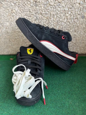 Svarta Ferrari sneakers med röd detalj - Snygga svarta sneakers i mocka med röda och vita detaljer samt Ferrari-logga på plösen. Skorna har svarta snören, röda öljetter och en vit vågig sidostrip. Yttersulan är svart och det följer med extra vita skosnören.