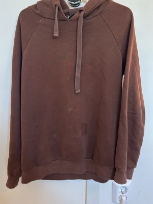 Brun hoodie med huva och dragsko - Mysig brun hoodie med huva och långa ärmar. Tröjan har ribbade muddar vid ärmslut och nederkant samt en klassisk dragsko i huvan. Perfekt för chill dagar eller när du vill ha något bekvämt och enkelt.