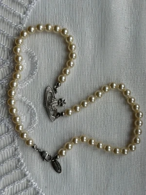 Vivienne westwood halsband - Snyggt halsband med glansiga vita pärlor och ett unikt silverfärgat hänge i form av en planet med gnistrande stenar. Halsbandet har ett klassiskt och trendigt uttryck, perfekt för att lyfta vilken outfit som helst. Låset är i metall och designen känns både modern och tidlös.