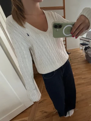 Vintage oversized vit stickad tröja - Vintage Kabelstickad oversized vit tröja från ralph lauren sport med V-ringning och lång ärm. Tröjan har ett diskret blått broderat märke på bröstet och är gjord i ett mjukt material. Klassisk och tidlös design som passar till mycket.