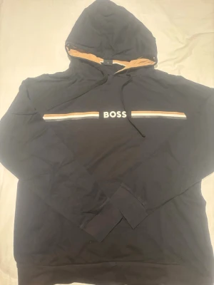 Svart hoodie från BOSS med huva - M men passar S bättre Snygg svart hoodie från BOSS med huva och dragsnören. Framtill finns BOSS-loggan tryckt mellan två ränder i vitt och beige. Insidan av huvan är beige för en schysst kontrast. Perfekt för en avslappnad och trendig look.
