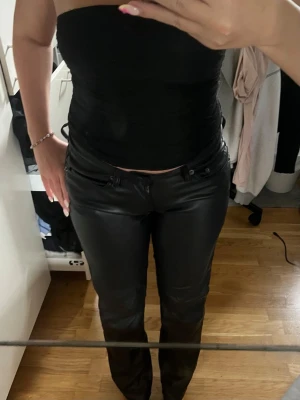 Svarta raka skinnbyxor - Säljer ett par svarta skinnbyxor med rak passform och klassisk midja. Byxorna har bälteshällor, knapp och dragkedja framtill. Perfekta för dig som vill ha en clean och edgy look.