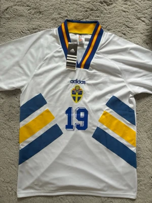 Sverige 94 borta tröja Adidas  - Vit svensk landslagströja från Adidas med blå och gula detaljer, nummer 19 och namnet ANDERSSON på ryggen. Tröjan har klassisk v-ringad krage med blå och gula ränder samt broderad svensk sköld på bröstet. Perfekt för sport och supporterhäng.