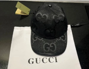 Svart Gucc* keps - Snygg svart keps från Gucc* med det klassiska GG-mönstret över hela kepsen. Tillverkad i ett strukturerat tyg med lätt glansig finish och böjd skärm. Perfekt accessoar för dig som vill ha en exklusiv och trendig look.
