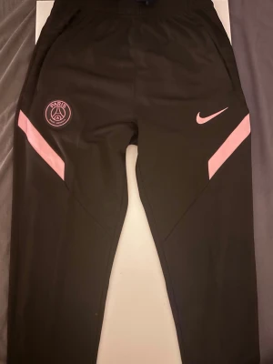 Svarta PSG träningsbyxor Nike - Säljer ett par svarta träningsbyxor från Nike med PSG-logga och rosa detaljer. Byxorna har elastisk midja och smal passform, samt rosa Nike Swoosh och Paris Saint-Germain-märke på framsidan. Perfekta för fotbollsträning eller chill.
