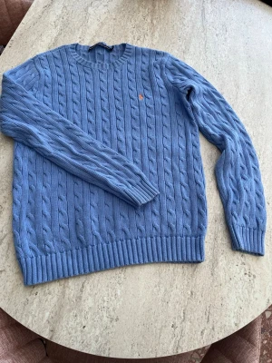 Blå kabelstickad tröja från Polo Ralph Lauren - Kabelstickad blå tröja från Polo Ralph Lauren med rund halsringning och orange broderad logga på bröstet. Tröjan har ribbade muddar vid ärmslut och nederkant och är tillverkad i mjukt material. Perfekt för en stilren och avslappnad look.