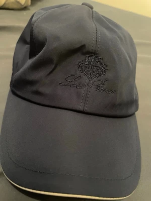 Loro piana keps - Snygg mörkblå keps från Loro Piana med broderad logga framtill. Kepsen har ett klassiskt böjt brätte och insidan är fodrad med rutigt tyg. Tillverkad i Italien och har Storm System-material som står emot väder och vind.