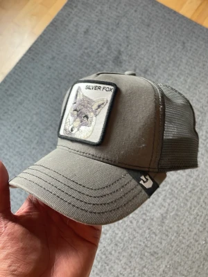 Grå keps med Silver Fox patch - Stilren grå keps med broderad patch framtill som föreställer en silverräv och texten 'SILVER FOX'. Kepsen har mesh på sidorna och justerbar snapback baktill. Perfekt för dig som gillar djurmotiv och streetwear.