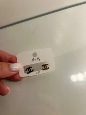 Guldiga örhängen med CC-logga - Säljer ett par små örhängen i metall med den klassiska CC-loggan i guld och svart. Örhängena har en stilren och trendig design som passar perfekt till alla tillfällen. Fästs enkelt med plupp på baksidan.