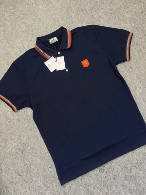Pikétröja  - Snygg marinblå pikétröja med orange och vita detaljer på krage och ärmslut. Tröjan har knappar framtill och ett orange broderat H-märke på bröstet. Perfekt för en stilren och sportig look.