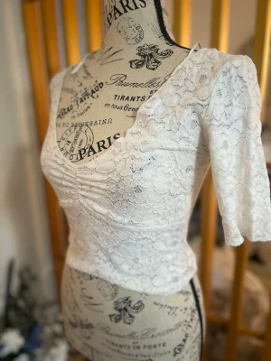 Vit spetsblus med v-ringning - Supersöt vit blus i spets med v-ringad hals och trekvartsärm. Blusen har fina rynkdetaljer framtill och är figurnära i modellen. Perfekt att styla med jeans eller kjol för en trendig look.