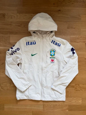 Vit Brasilien vindjacka med huva - Snygg vit vindjacka med huva och dragkedja framtill. Jackan har Brasilien-logga, grönt Nike-märke och blåa tryck med 'Itaú' och 'vivo' på bröst och ärmar. Lätt material, perfekt för blåsiga dagar. Sportig look med fickor och justerbara ärmslut.