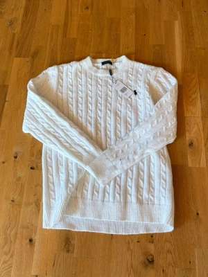 Ralph lauren Tröja - Kabelstickad vit tröja från Polo Ralph Lauren med rund halsringning och svart broderad logga på bröstet. Tröjan har ribbade muddar vid ärmslut och nederkant och är tillverkad i mjukt material. Perfekt för en klassisk och stilren look.