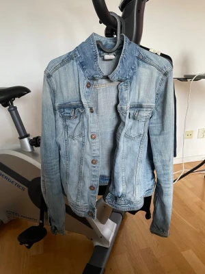 Ljusblå klassisk jeansjacka - Snygg ljusblå jeansjacka med bruna knappar och två bröstfickor med lock. Jackan har en klassisk krage och raka ärmar. Perfekt att slänga över en hoodie eller t-shirt för en avslappnad look.