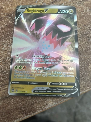Pokémonkort Regidrago V - Säljer ett Pokémonkort av typen Regidrago V med holografisk yta. Kortet har färger i svart, rosa och gult med cool drakdesign och tydliga attacker som Celestial Roar och Dragon Laser. Perfekt för samlare eller Pokémonfans som vill utöka sin kortlek.