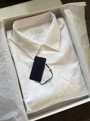 Prada Popeline Shirt – Helt ny med tags & box - Prada Popeline Shirt i vitt, storlek XXL. Helt ny och oanvänd med originaletiketter och originalbox. 100 % äkta. Säljs eftersom den inte kommer till användning. Kan skickas eller hämtas. Hör av dig vid frågor eller om du vill ha fler bilder. Pris: 5000 kr