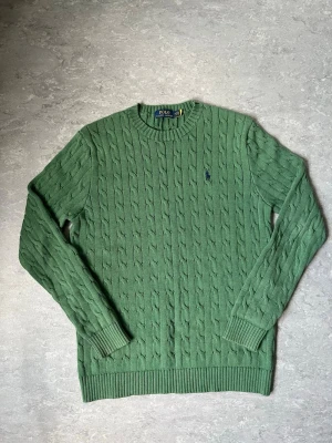 Grön kabelstickad tröja Polo Ralph Lauren - Snygg grön kabelstickad tröja från Polo Ralph Lauren med klassisk rund halsringning och ribbade muddar. Tröjan har det ikoniska broderade logotypen på bröstet och är tillverkad i mjukt material som känns skönt mot huden. Perfekt för dig som gillar preppy stil.