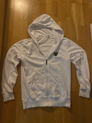 Vit hoodie från Comme des Garçons Play - Säljer en vit hoodie från Comme des Garçons Play i storlek S. Hoodien har dragkedja, fickor framtill och den ikoniska svarta hjärtloggan på bröstet. Tillverkad i mjukt bomullsmaterial och har en klassisk huva. Perfekt för en clean och stilren look.