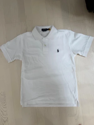 Vit pikétröja från Polo Ralph Lauren - Klassisk vit pikétröja från Polo Ralph Lauren med broderad logga på bröstet. Tröjan har korta ärmar, krage och knappar framtill. Modellen är custom slim fit och passar perfekt till en stilren look. Har Andvänds 2 gånger. Storlek S