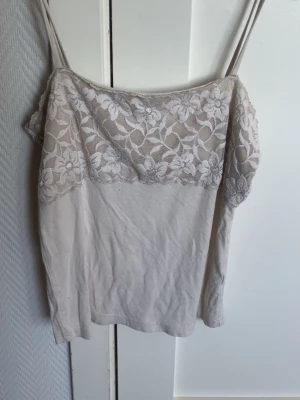 Beige linnetopp med spetsdetaljer - Säljer en ljusbeige linnetopp med tunna axelband och vacker spets upptill. Toppen har ett blommigt spetsmönster över bysten och är perfekt för sommaren. Materialet känns mjukt och lätt, och toppen är ärmlös för en luftig känsla.