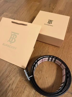 Burberry svart bälte - Snyggt svart bälte från Burberry med klassiskt rutigt mönster på insidan och en stor metallspänne med Burberry-logga. Bältet är gjort i skinn och har en stilren design som passar till många olika outfits.