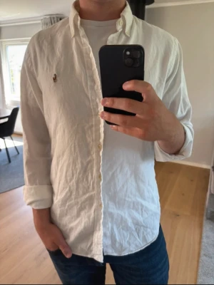 Vit skjorta med knappkrage - Klassisk vit skjorta med knappkrage och broderad logga på bröstet. Skjortan har långa ärmar och knappar framtill. Perfekt till jeans eller chinos för en clean och stilren look.