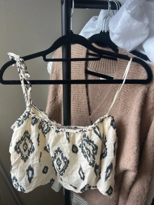 Mönstrad linnetopp från Zara - Säljer en krämvit linnetopp från Zara med svart grafiskt mönster. Toppen har smala axelband, varav ett är flätat, och ett luftigt, croppat snitt. Perfekt för varma dagar och enkel att styla med jeans eller kjol.
