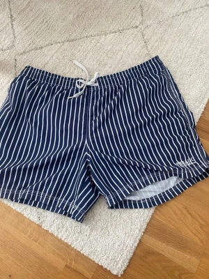 Randiga badbyxor från Sebago - Snygga marinblå badbyxor från Sebago med broderad logga på benet. Klassisk modell med dragsko i midjan och innerbyxa i mesh.