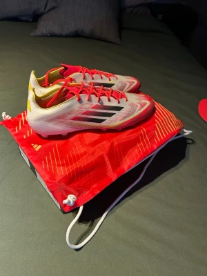 Adidas F50 - Snygga fotbollsskor från Adidas i vitt med röda och gula detaljer samt svarta ränder på sidan. Skorna har röda snören och en låg profil för bra bollkontroll. Ovandelen är tillverkad i ett lätt syntetmaterial som ger bra känsla på planen.  Box , sulor, påse och kvitto ingår