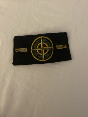 Stone Island tygmärke svart/gult - Säljer ett klassiskt Stone Island tygmärke i svart med gul brodyr och kompasslogga. Märket har två gula knapphål på sidorna och är perfekt att fästa på jackor eller tröjor för en streetwear-look.